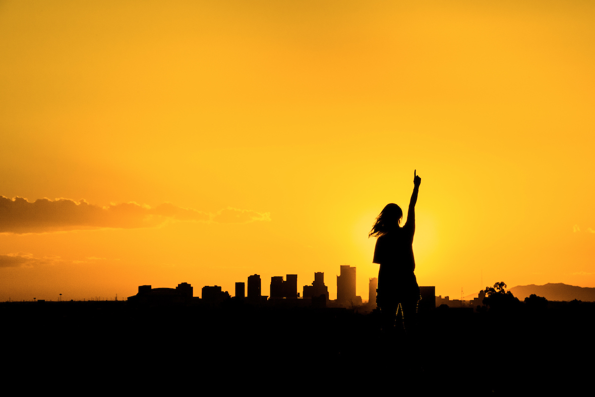 ASU student #1 A person in shadow stands at sunset in front of a shadowed city skyline making the "number one" gesture with her left hand.