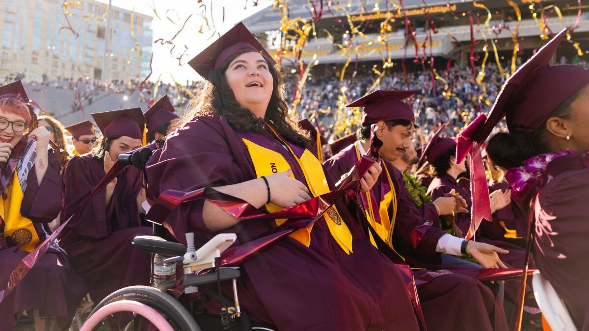 A graduate in maroon and gold regalia sits in a mobility device while watching maroon and gold streamers during University Commencement.