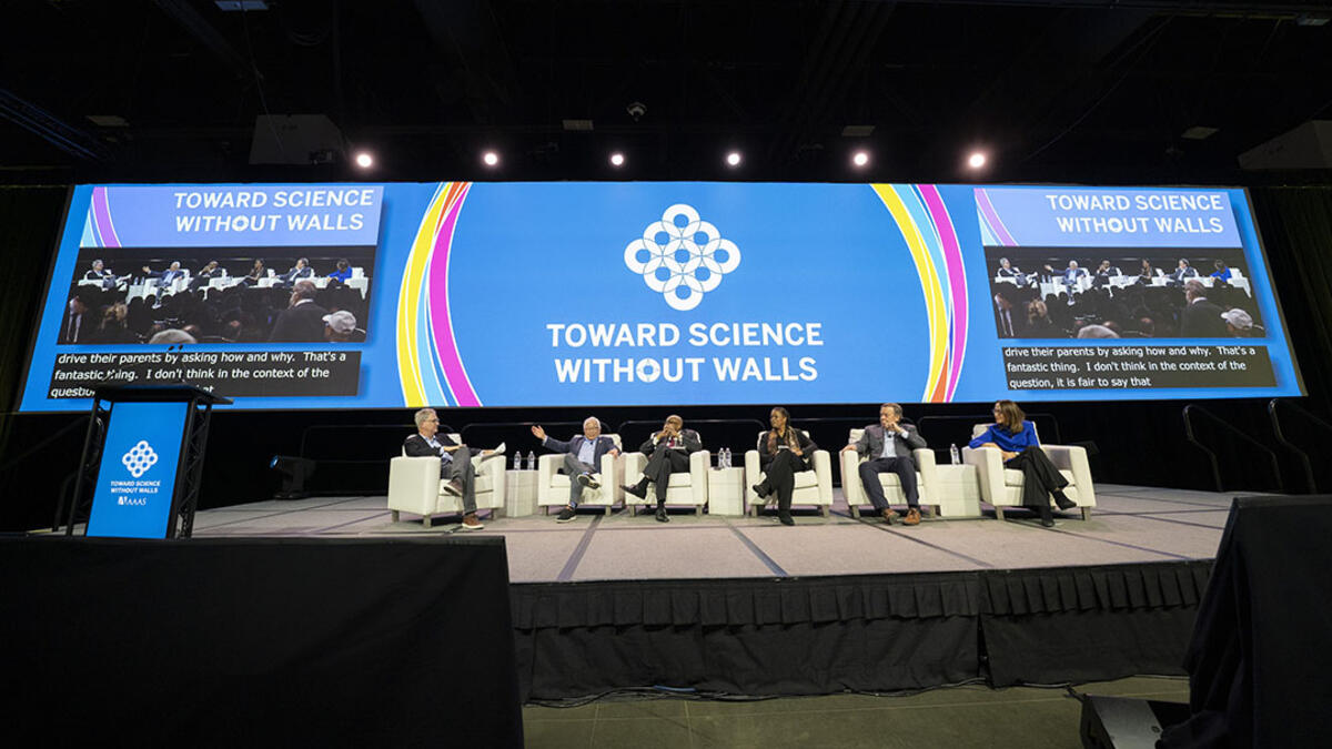 Six people sit in chairs on a stage and a large LED screen discussing higher education and science before a live audience.