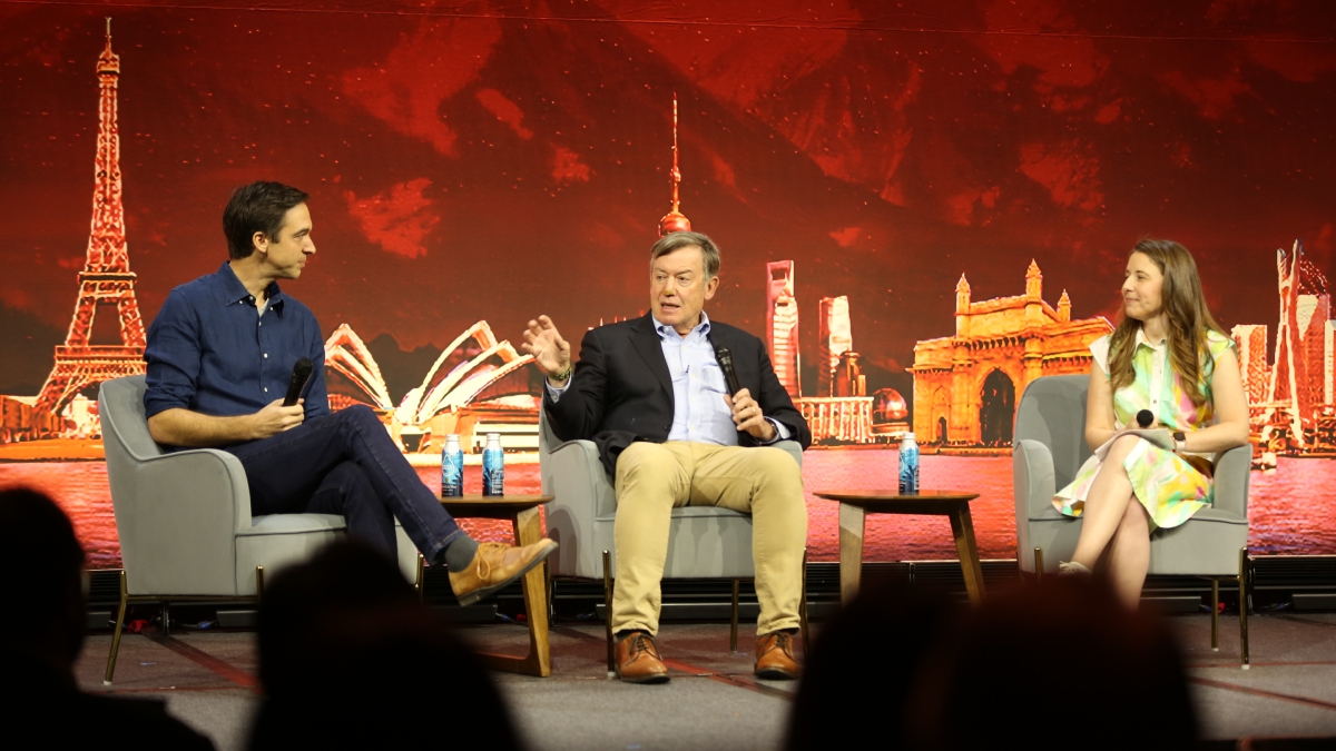Three people sit on-stage un front of a backdrop featuring famous buildings from around the world while discussing learning through games.