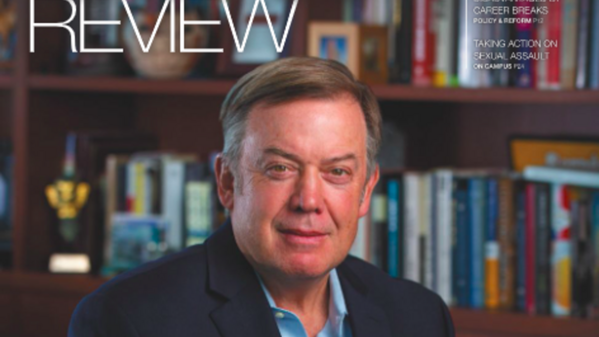 A adult wearing a navy jacket and light blue shirt sits in front of a bookcase. 