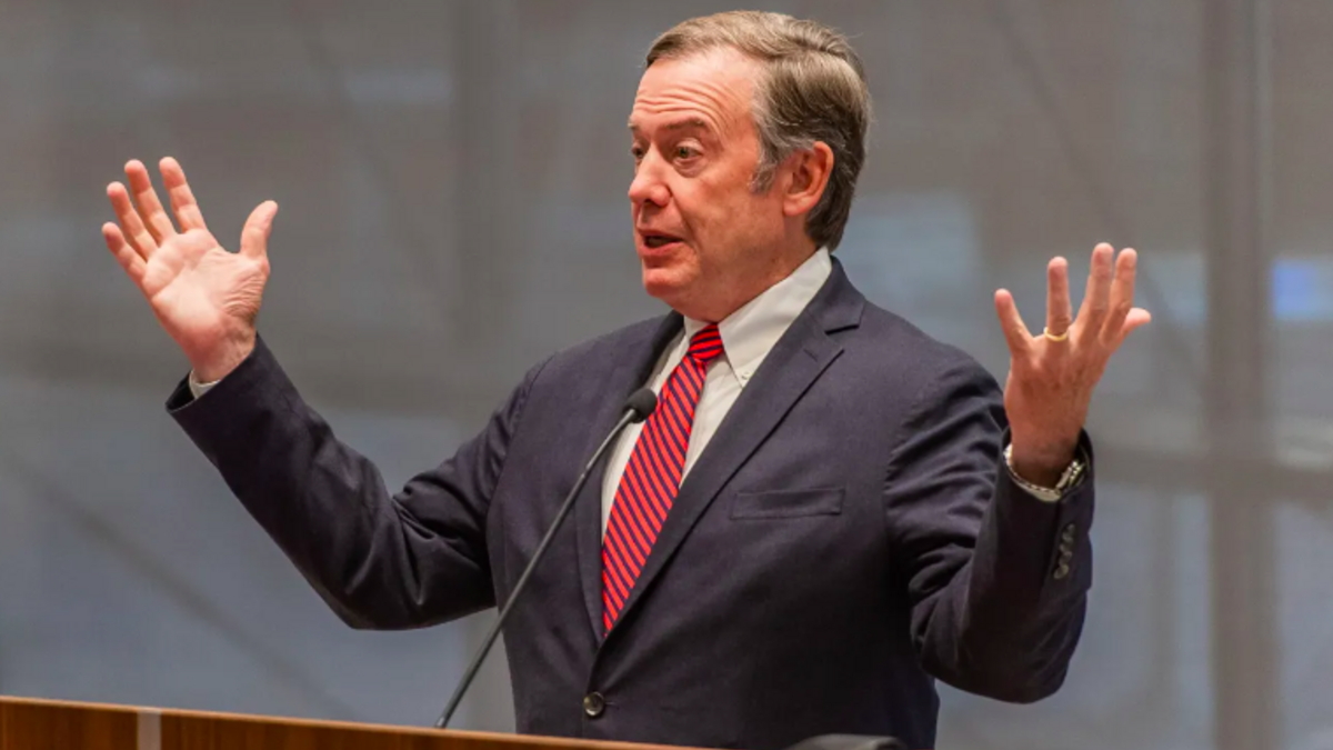 A person wearing a blue jacket and red striped tie lifts his hands while speaking at a podium in the ASU Center for Law and Society. 