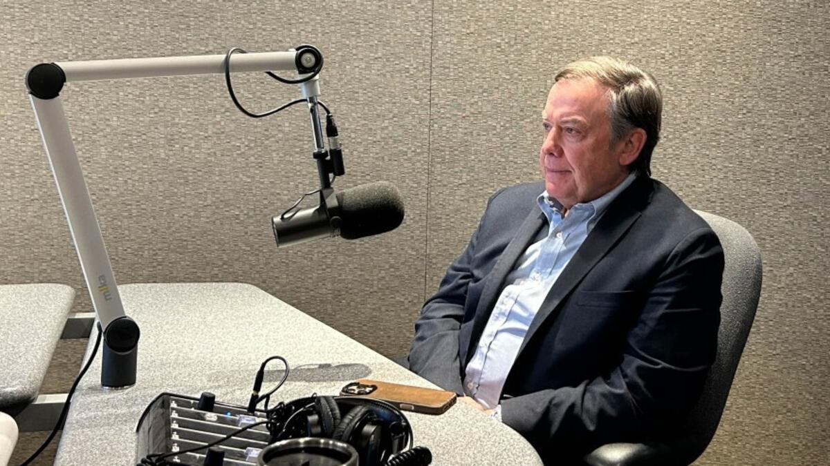 A person in a blue shirt and blazer sits in a radio studio preparing for an interview on federal changes to American higher education.