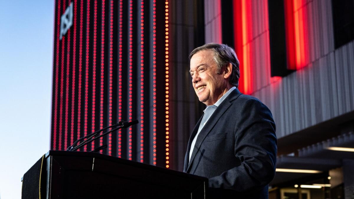 A person stands at a podium smiling with excitement in front of a crimson-lit new building at its grand opening.