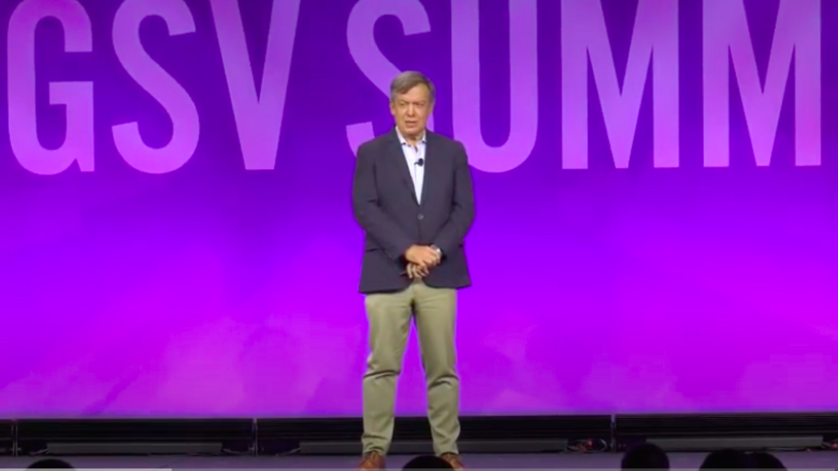 A middle-aged man stands on a stage in beige slacks and a blue blazer in front of a backdrop that reads "GSV Summit."